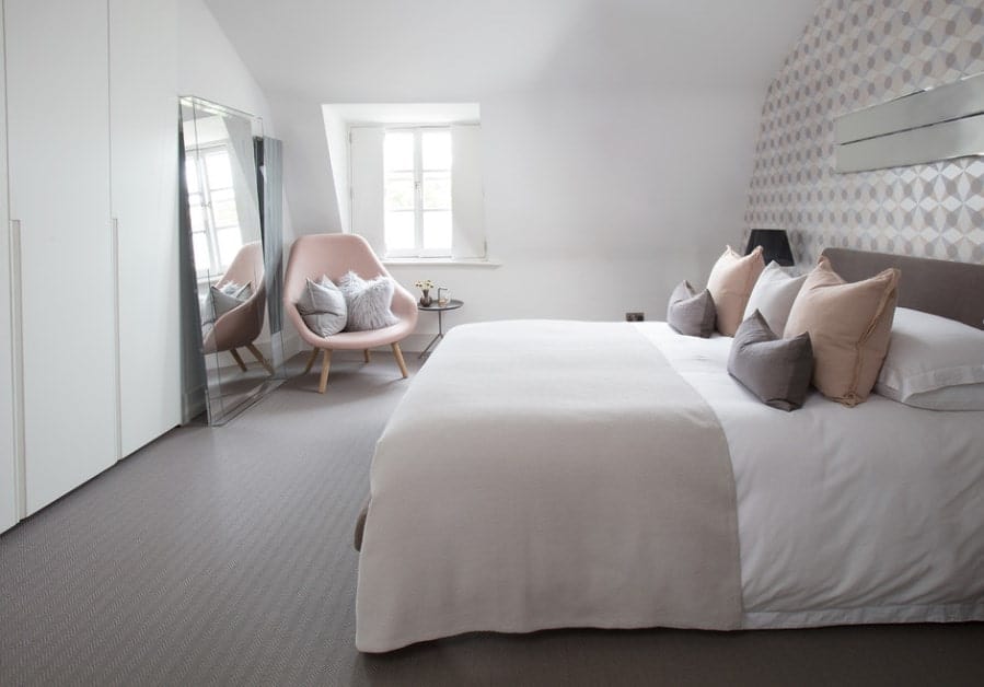 Classy Scandinavian-Style primary bedroom featuring gray rug flooring and a stylish wall. Thereโs a cute pink chair on the corner near the window.