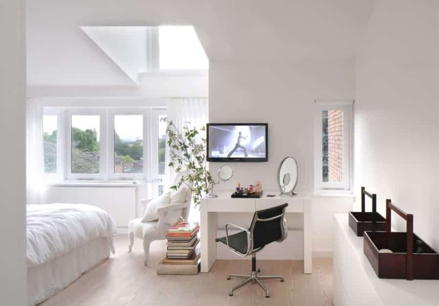 A classy Scandinavian-Style primary bedroom featuring white walls and ceiling along with white window curtains and a makeup desk with a TV on the wall.