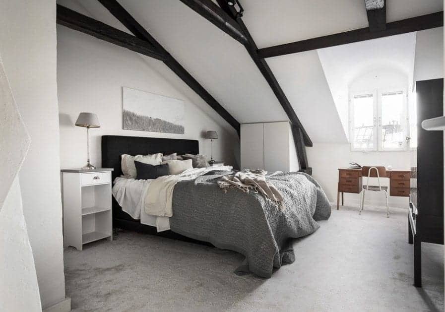 A stylish Scandinavian-style primary bedroom offering an elegant black bed frame set on the gray carpet flooring. The espresso-finished beams on the ceiling add style and elegance to the room.