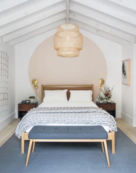 This is a cozy alcove Scandinavian-Style bedroom with a pedant light covered in woven wicker hanging from the white cathedral ceiling that has exposed wooden beams. The wooden headboard of the bed is against the white wall painted with a huge beige circle that matches the wall-mounted painting.