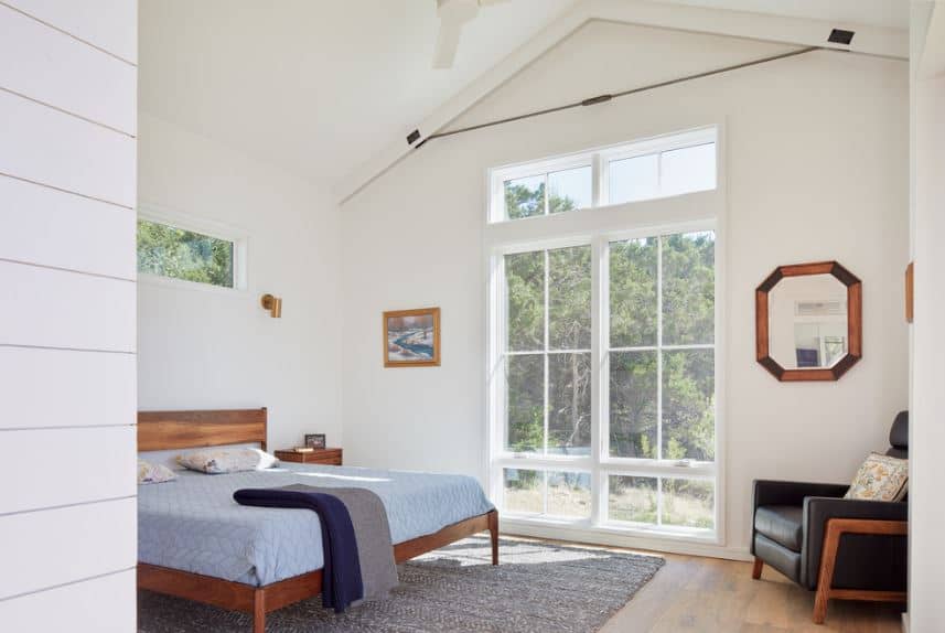 This is an airy Scandinavian-Style room with an abundance of natural light coming from the tall glass window that almost goes up to the white cathedral ceiling. This illuminates the wooden traditional bed that has light blue sheets contrasting the gray area rug over the hardwood flooring.