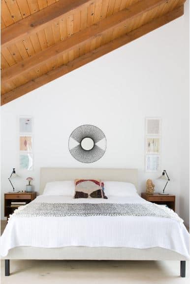 The shiplap wooden ceiling of this Scandinavian-Style bedroom has exposed wooden beams that contrast with the white walls and white flooring. The light gray hue of the headboard molds into the white wall above it and accented with a circular artwork.