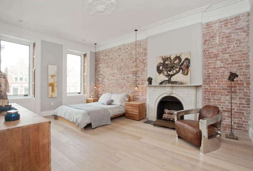 This wide and airy Scandinavian-Style bedroom has hardwood flooring, white ceiling, and red brick walls. The simple wooden platform bed has bedside tables that were made from crates. Next to it is an elegant fireplace with white stone fitting and a colorful painting above it.