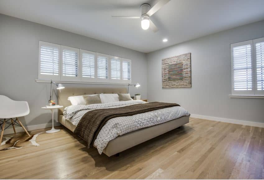 The hardwood flooring is illuminated by the pin lights and ceiling fan with semi-flush light mounted on the white ceiling. There is a row of small shuttered windows above the gray cushioned headboard that is flanked by modern table lamps on white bedside tables.