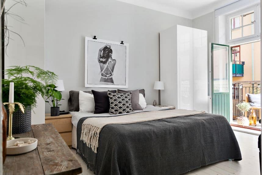 The natural light coming in from the French glass doors and windows above it illuminate the white walls, ceiling and the sleek white cabinet beside the bed. There is a lovely framed photo of a woman mounted above the bed with gray sheets.