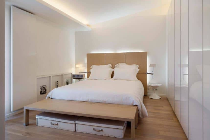The white shed ceiling of this Scandinavian-Style bedroom is paired by two distinct white walls with different designs. One wall has a sleek and shiny finish with stripes while the wall across it has wall-mounted artworks and overlaps with the ceiling. This is a nice background for the wooden platform bed.