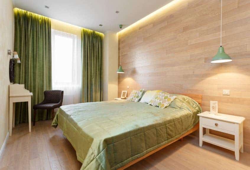 The chic greenish sheets of the traditional wooden bed match well with the green curtains of the tall windows. These green hues are a perfect contrast for the white ceiling, wooden walls, and hardwood flooring.