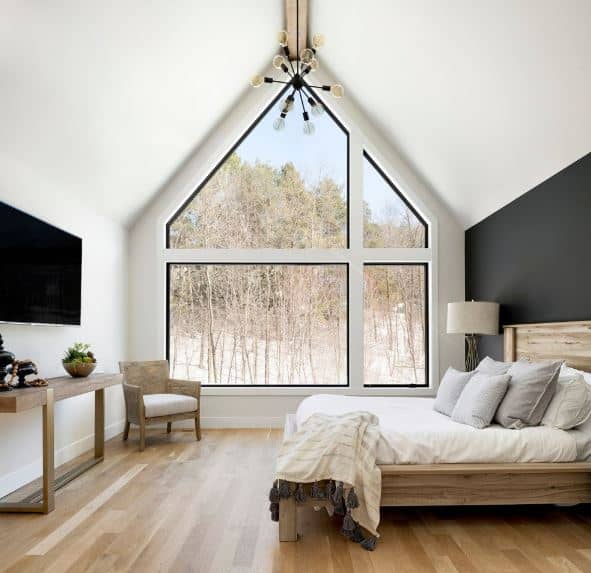 The massive glass window of this Scandinavian-Style bedroom follows the lay of the high white cathedral ceiling with a single exposed wooden beam in the middle that supports a modern chandelier. The wall behind the wooden traditional bed is painted dark gray contrasting the other walls.