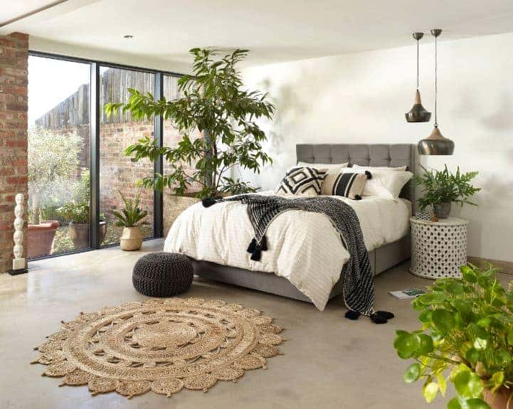This bedroom has white walls and a white ceiling that serve as a nice background for the plant accents that match with the plants outside the glass floor-to-ceiling window. This contrasts well with the dark gray cushions of the traditional bed.