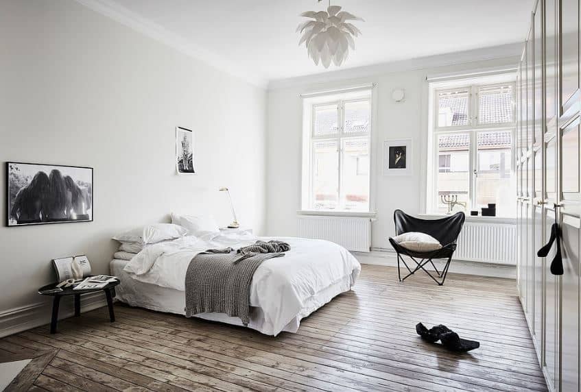 This is a bright and airy Scandinavian-Style bedroom with hardwood floors illuminated by the pair of tall windows dominating one wall. The white sheets of the cottage-style bed are contrasted with a dark wooden bedside table and wall-mounted black and white photo.