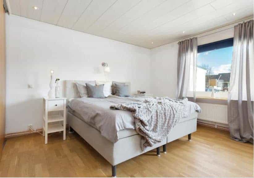 The light gray platform bed stands out against the white walls of this Scandinavian-Style bedroom. The wall above the bed has a wall-mounted modern lamp for late-night reading. This matches with the white wooden bedside tables.