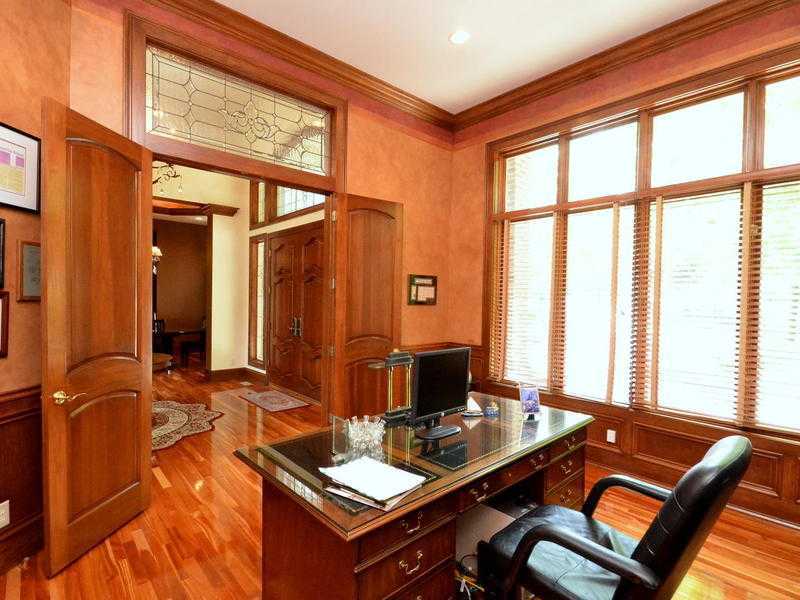 Warm home office boasts a rich hardwood flooring and glass windows that bring natural light in. It includes a wooden desk paired with a black leather chair.
