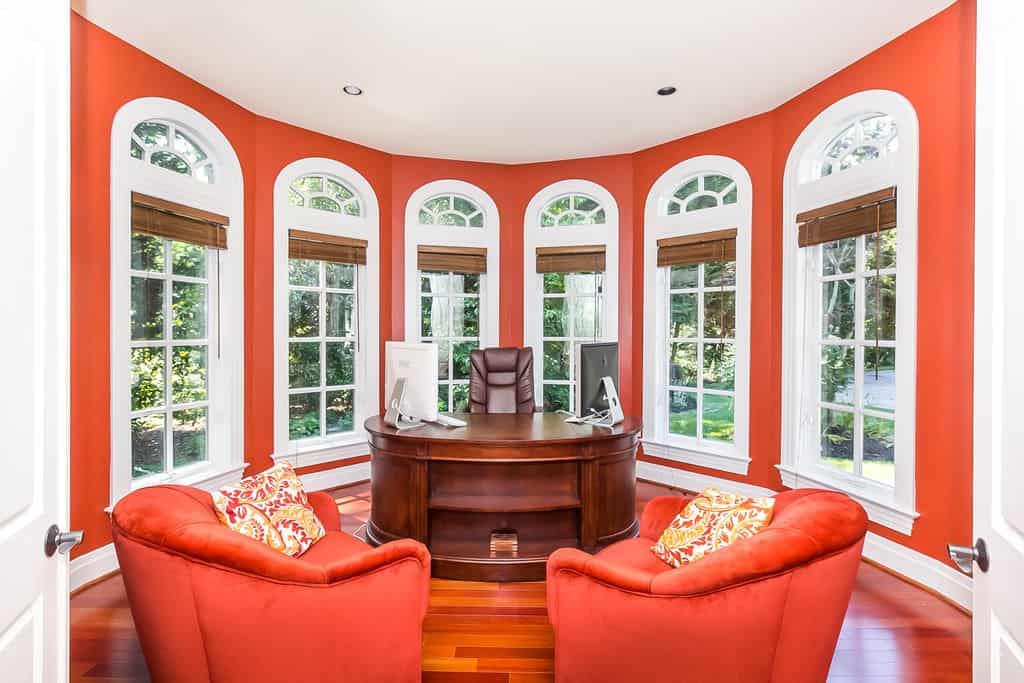 Curved red home office surrounded with full height arched windows covered with wicker blinds. It has an oval desk paired with a brown leather chair and red round back chairs.