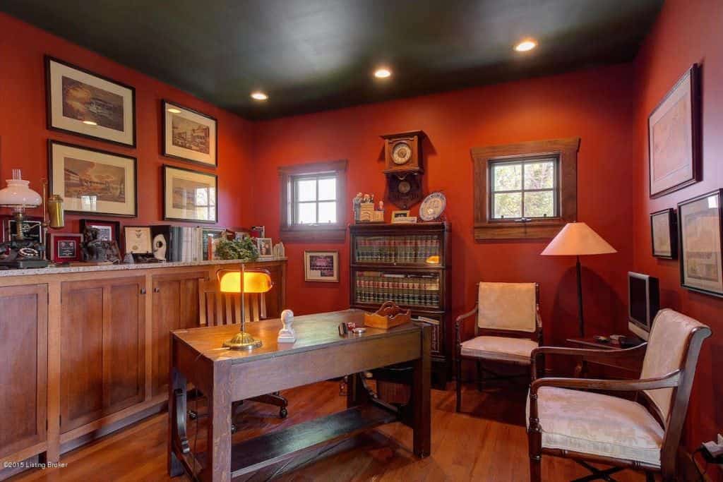 Red home office with hardwood flooring and black ceiling fixed with recessed lighting. It has a wooden desk with a matching chair and storage cabinet topped with books and frames.