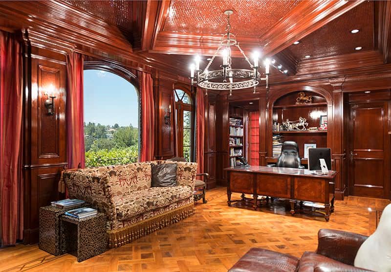 Sophisticated home office boasts redwood walls and tray ceiling with a hanging wrought iron chandelier. It has a classy seating area by the window adjacent to the wooden desk paired with a black leather office chair.