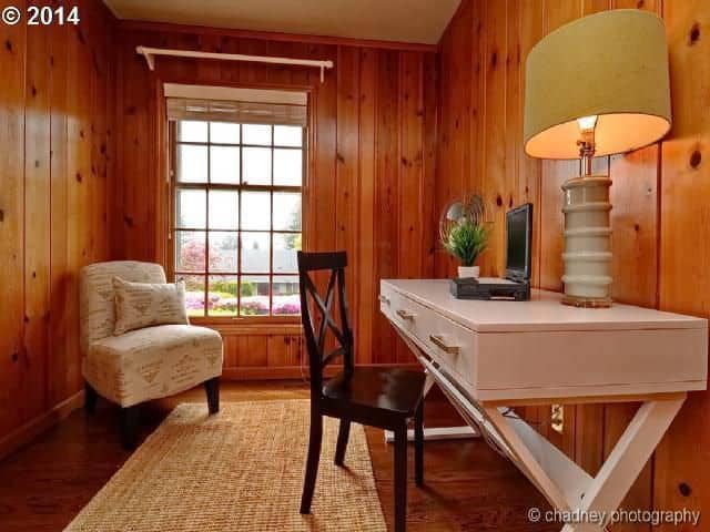An all wood home office features wood plank walls fitted with a picture window. It has a white comfy chair along with a black wooden chair that sits at a white office desk topped with a table lamp.