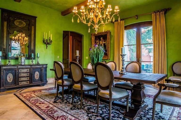 Green dining area lighted by a lovely chandelier that hung over a wooden dining table with round back chairs. It sits on a vintage rug over tiled flooring.
