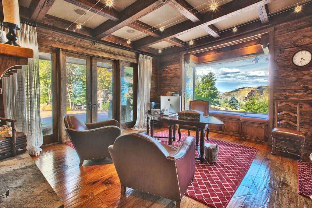 Cozy home office with wood coffered ceiling and hardwood flooring topped by lovely red patterned rugs. It has glazed doors and a window that overlooks a stunning mountain view.