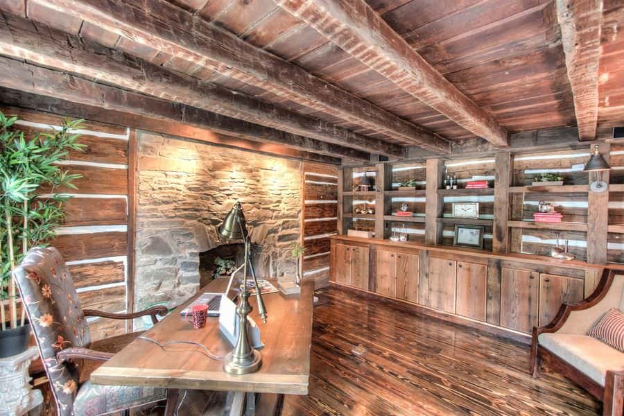 Rustic home office furnished with a wooden table and floral chair along with a beige lounge chair that sits in front of the built-in shelves. It includes a stone fireplace lined with wooden panels. 