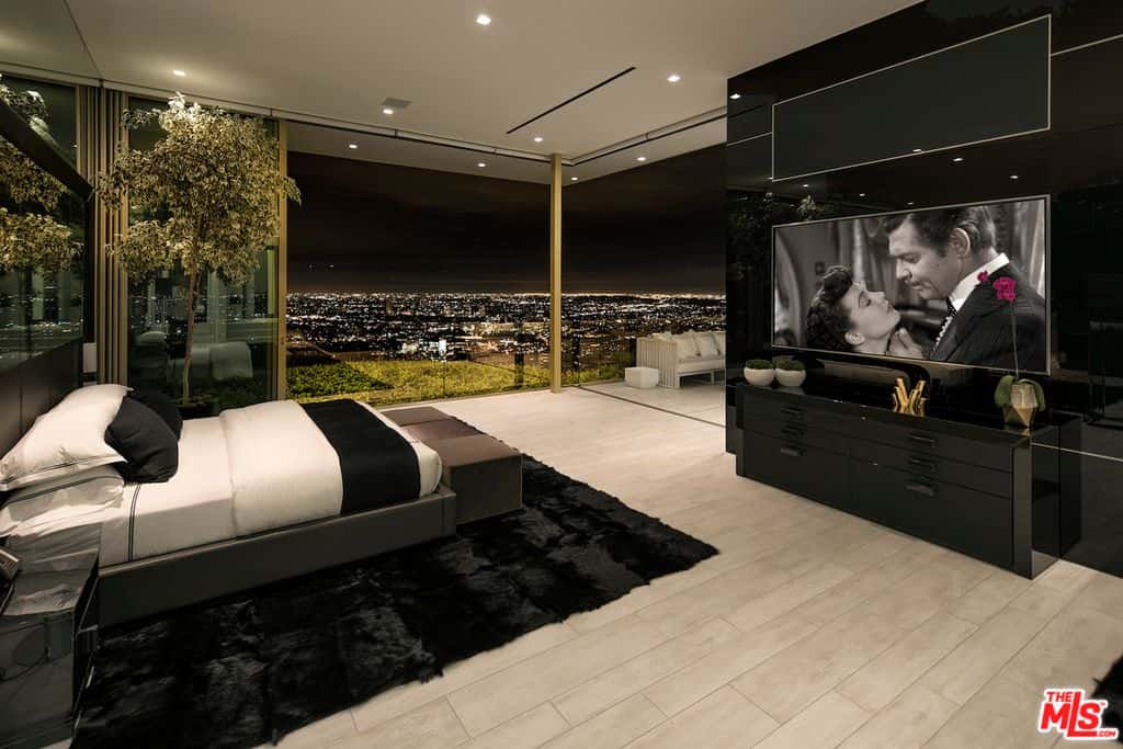 Luxurious primary bedroom features a bed with bench that sits on a black shaggy carpet and facing the absolute black polished wall where the television is mounted. It has an astounding city view from the frameless glass windows.