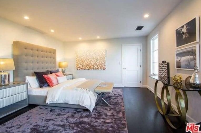 A spacious primary bedroom with a luxurious bed, a stylish purple rug and white walls, lighted by recessed ceiling lights.