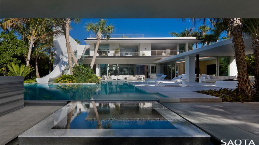 This home features an infinity pool with a slide. Multiple lounging spaces are on the side.