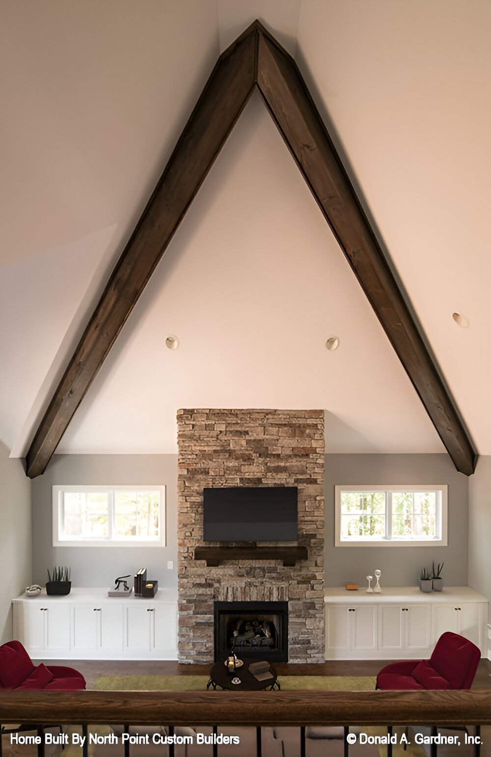 The great room has red armchairs, a stone fireplace with a TV on top, and a high cathedral ceiling framed with rustic beams.