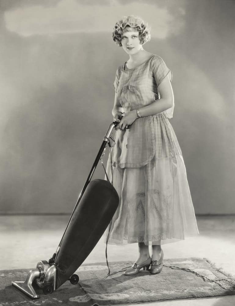 Lady from a previous era holding one of the earliest versions of a vacuum cleaner.
