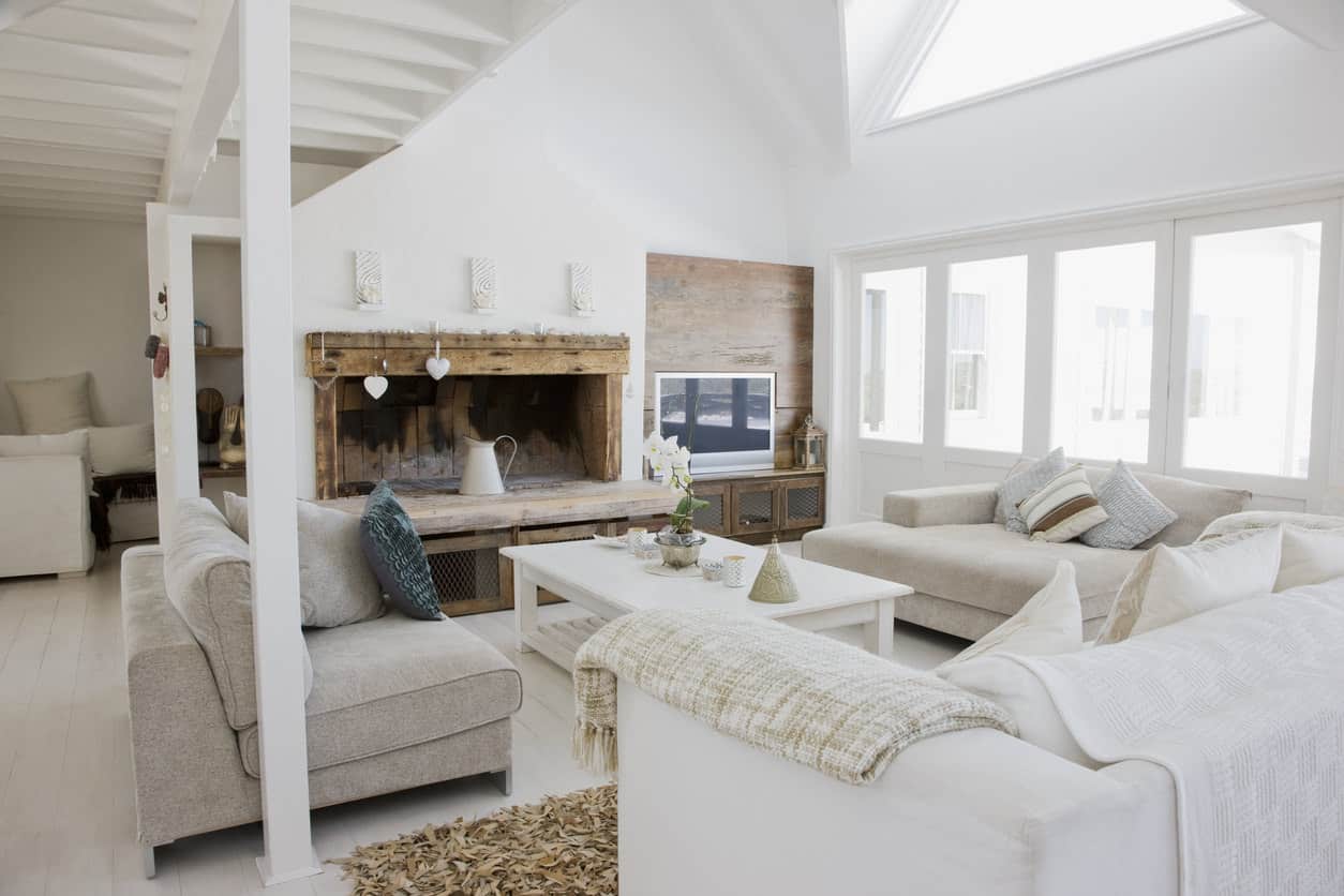 Here's a light rustic living room with towering white ceiling and distressed beamed fireplace mantel. The light beige sofas tie in the distressed wood with the otherwise all white space.