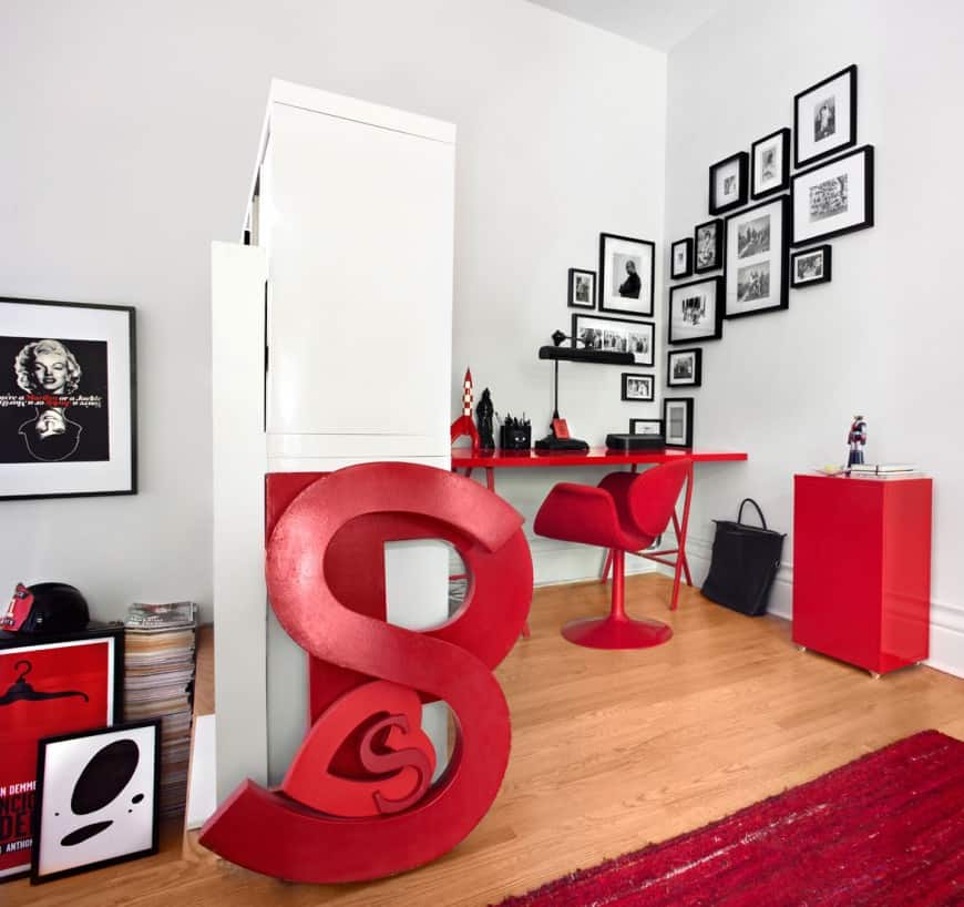Gorgeous home office designed with huge red letter decors and gallery frames mounted on the light gray wall. It has a red office desk and chair over hardwood flooring topped by a red rug.