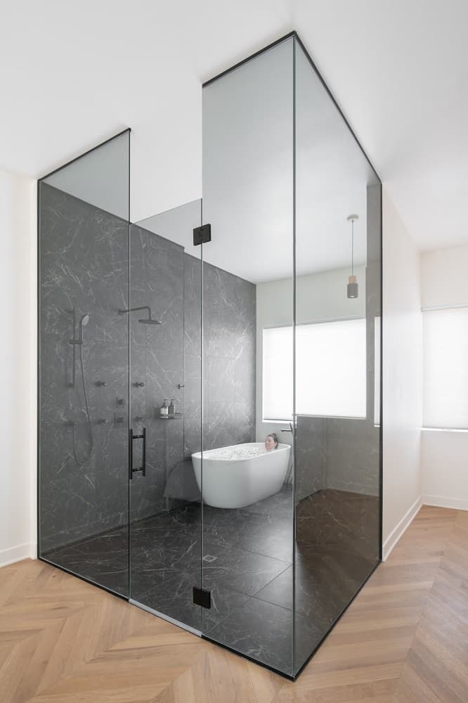 This is a close look at the bathroom that has glass walls on two sides and black marble on one side as well as its flooring that makes the white freestanding bathtub at the far corner stand out.