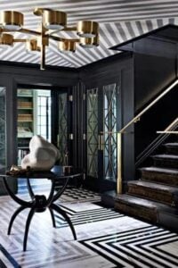 Stunning black and white foyer with striped tile flooring and matching ceiling. The doors are an elegant glass with brass details and a grand staircase leading up to the main house, is absolutely stunning with those gold railings.
