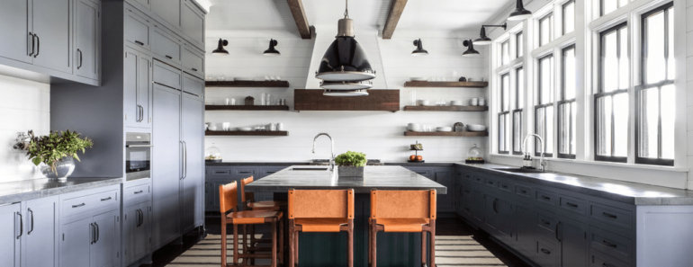 Spacious farmhouse kitchen with blue cabinetry, wood beams against white ceiling and rustic open-faced floating shelves by chango & co.