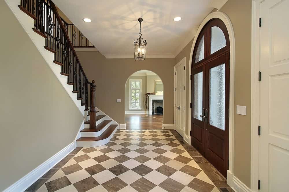 This classy foyer features a checker flooring, gray walls and vinyl staircase steps lighted by a stunning pendant light.