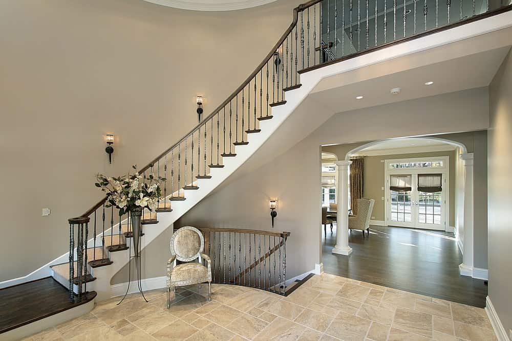 Large foyer featuring a glamorous staircase surrounded by gray walls and lighted by wall lights.