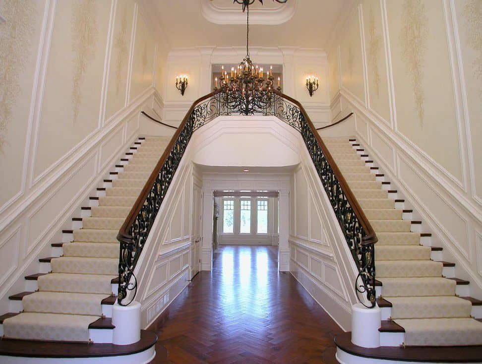 Large foyer with amazing staircases and hardwood flooring. The staircases' steps are topped by elegant rugs. The area is lighted by a grand chandelier.