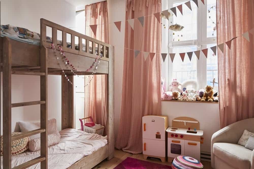 This beautiful girlโs bedroom has cute salmon-colored curtains that pair well with the beige walls. These serve as a pleasing background for the for the wooden bunk bed that matches the shelf for the toys under the window by the cushioned arm chair.