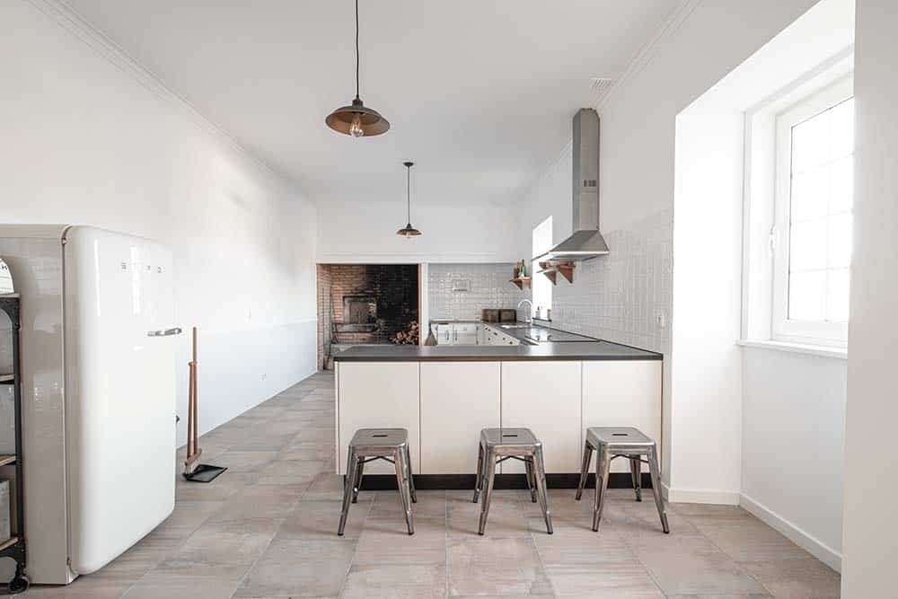 This is a full view of the simple kitchen with white cabinetry to its L-shaped peninsula with white subway tiles that blend with the white walls and white ceiling. These are then complemented by the rustic fire-burning oven on the far side with brick walls.