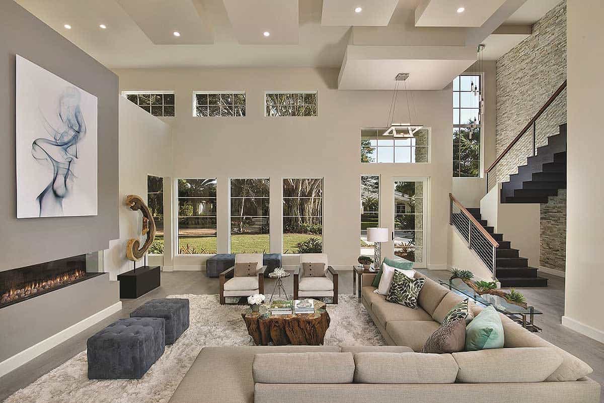 The high ceiling of this contemporary living room features recessed lighting and a dropped down pendant fixture. Each of the three walls in the massive space gets an entirely different treatment: one side wall is brick, a second side wall hosts a long fireplace and abstract wall art, while a third wall looks out into the backyard through a series of large windows. The seating area includes a large sectional sofa with chairs on an area rug, a coffee table made out of the section of a tree and two ottomans. The extreme modernity of the room's design doesn't give up its Mediterranean influences easily but the brick wall, neutral color palette and reliance on natural lighting all make conceptual connections.