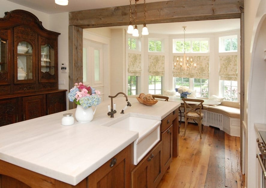 This kitchen features white walls and hardwood flooring along with a center island equipped with white smooth countertop lighted by pendant lights.