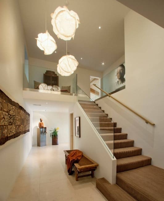 A modern house featuring an L-shape staircase with brown carpet floors and glass railings, lighted by a gorgeous pendant lighting.