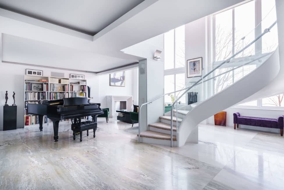 Large formal living space with a cozy sofa set near the fireplace, a large bookshelf near the grand piano and a stunning staircase with carpet floors and glass railings.