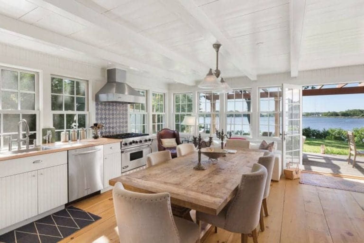 This single wall kitchen offers a wooden dining table set with classy chairs lighted by pendant lighting set on a white ceiling with beams.