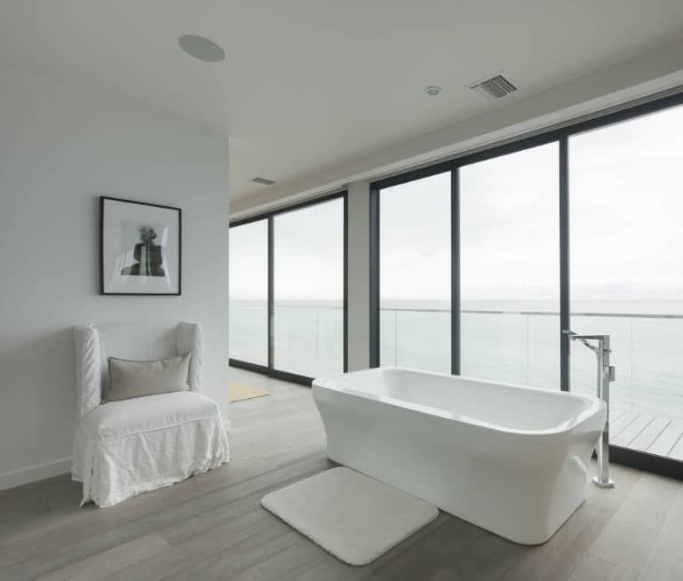 Large primary bathroom featuring gray walls and hardwood flooring, along with glass windows overlooking the stunning outdoor views. It also offers a freestanding tub and a white chair on the side.