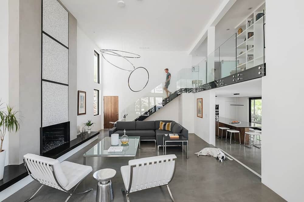 This is a full view of the modern living room that has a large white wall that houses the fireplace across from the black L-shaped sofa and the white cushioned chairs paired with a glass-top coffee table.