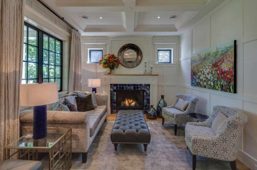 A round mirror placed above a blue brick fireplace creates a dramatic focal point in this craftsman living room. It has white panelled walls complementing the white coffered ceiling.