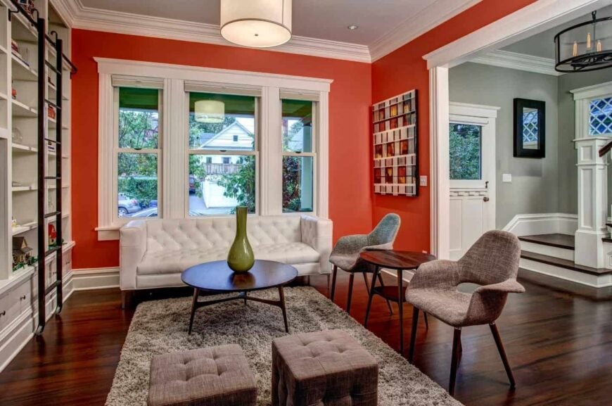 Vibrant red living room features white built-in bookshelves facing the white tufted sofa and brown chairs and stools. A wooden coffee table sits in the middle over a brown shaggy rug.