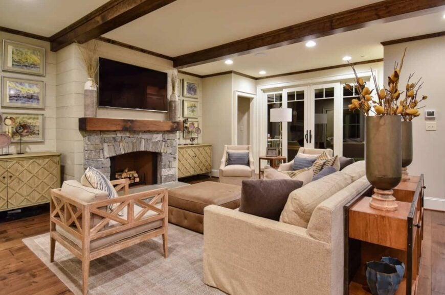 Gorgeous living room featuring a stone fireplace lined with a rustic wood that matches the exposed wood beams on the ceiling. It is fixed on a shiplap wall between wooden cabinets styled with frames and decors.