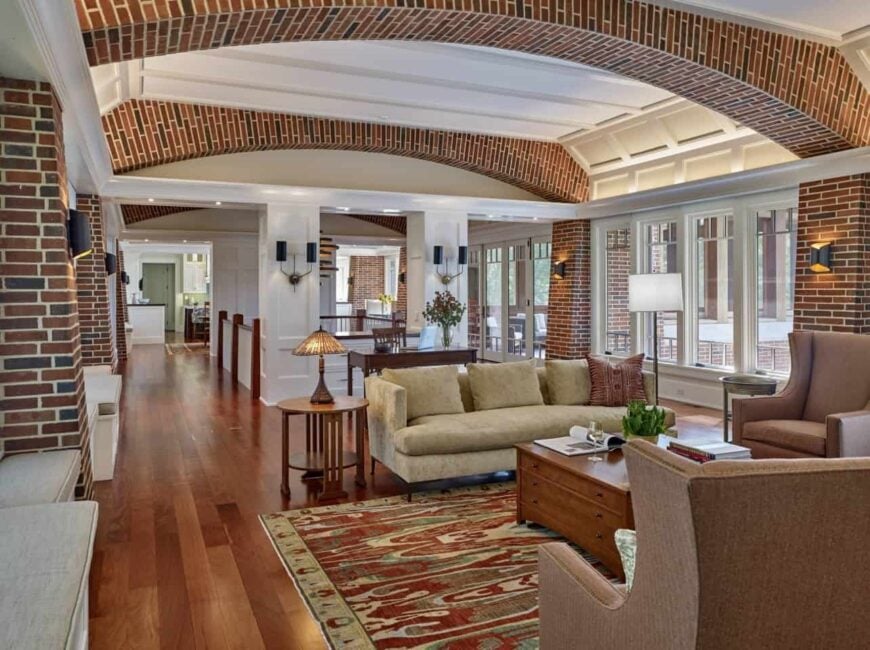 Magnificent living room accented with brick arches on the ceiling and pillars lighted by black wall sconces. It has a wooden center table surrounded with an upholstered sofa and brown wingback armchairs.