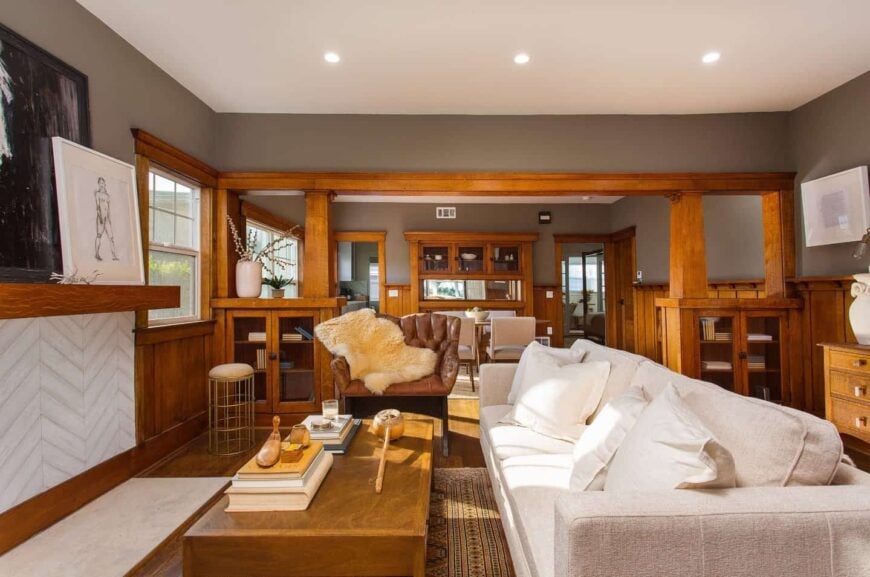 Craftsman living room features gray walls above wood wainscoting and white ceiling with recessed lighting. It has a long white sectional sofa paired with a leather tufted chair accented by a fur throw blanket.